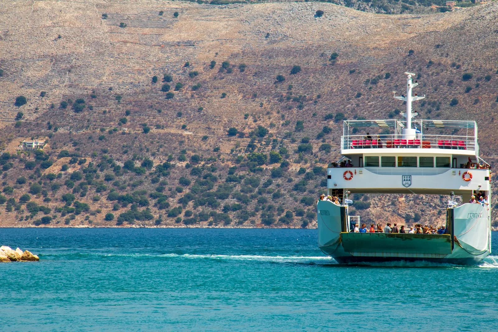 Un ferry chargé de passagers quitte le port d'une île grecque, ses collines arides plongeant dans une mer turquoise