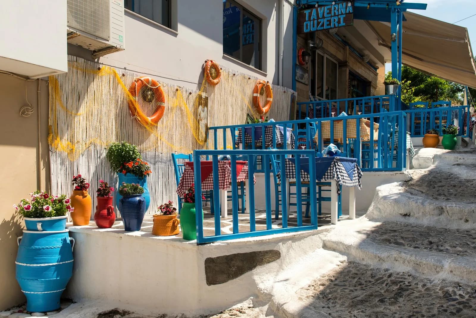 Terrasse d'une taverne grecque aux chaises bleues et nappes à carreaux, en bord de ruelle blanchie à la chaux
