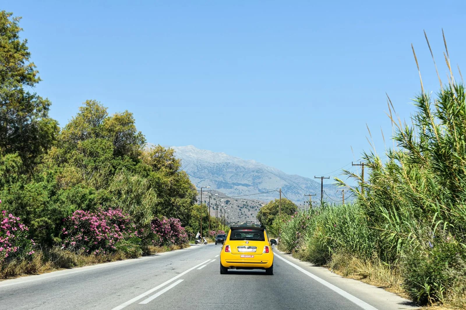 Petite voiture jaune immatriculée en Grèce sur une route bordée de lauriers-roses, montagnes de Crète au fond