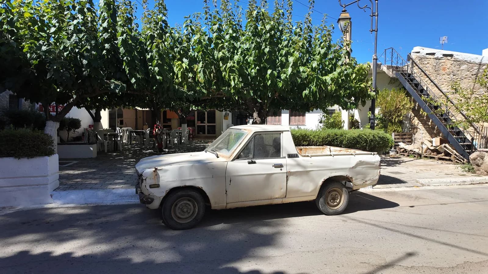 Vieux pick-up blanc cabossé garé sous les mûriers d'une place de village en Crète