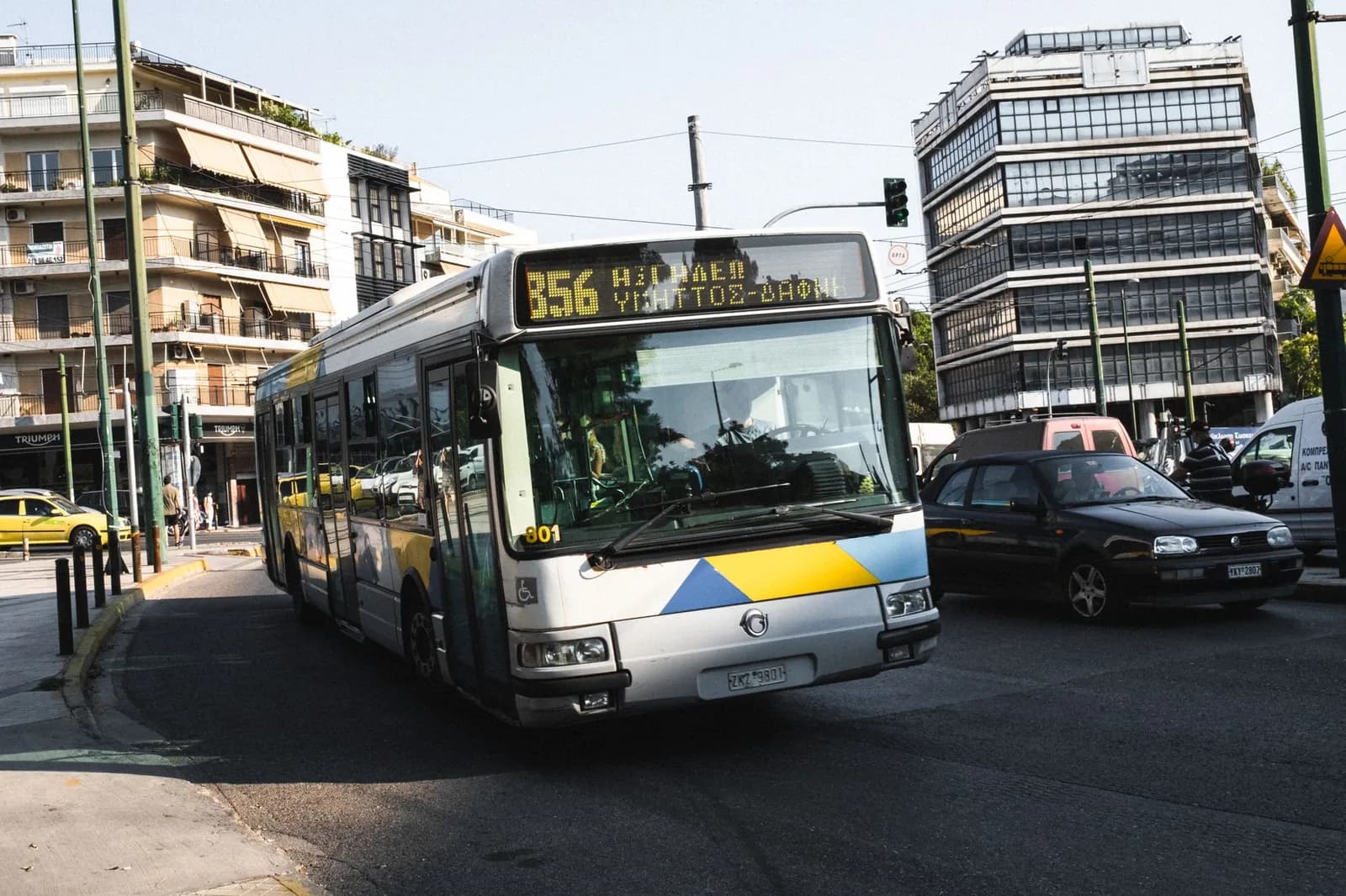 Un bus urbain jaune et bleu de la ligne 856 tourne dans une rue d'Athènes, un taxi jaune à l'arrière-plan