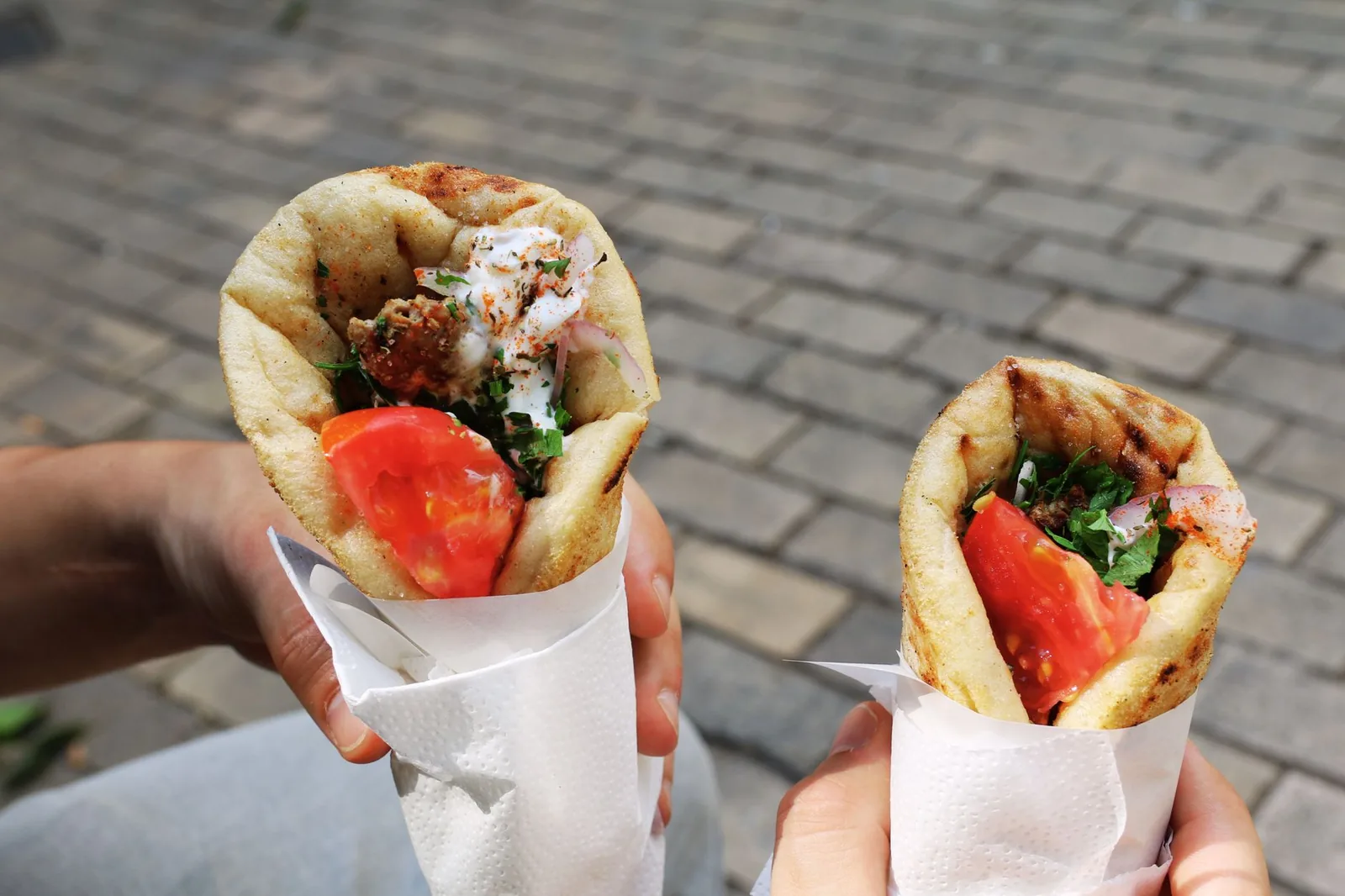 Deux pita gyros grecques garnies de tomate, oignon et sauce, tenues à la main dans une rue pavée