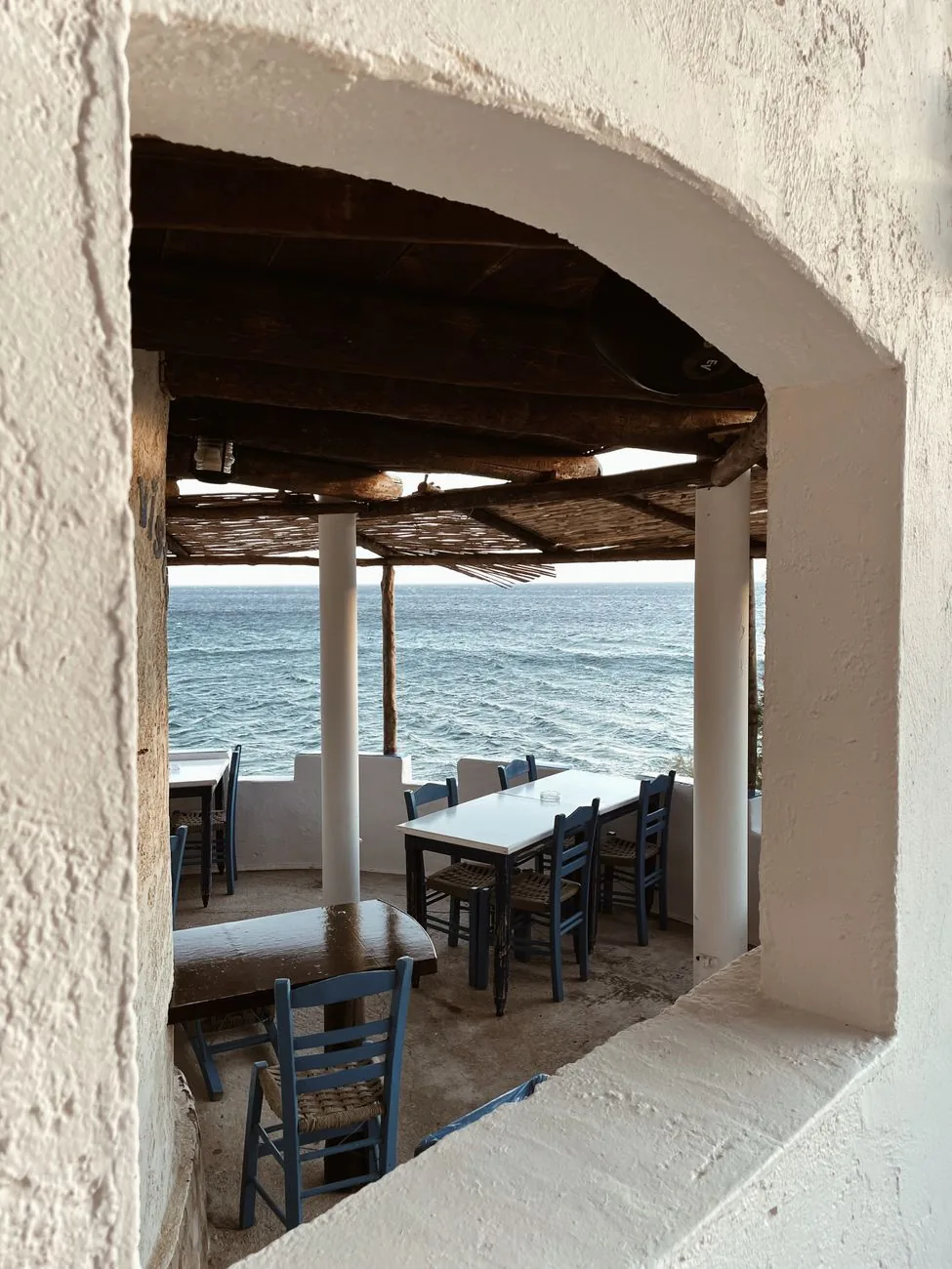 Terrasse de taverne grecque aux chaises bleues et à la pergola de bambou, vue sur la mer Égée depuis une fenêtre blanchie à la chaux