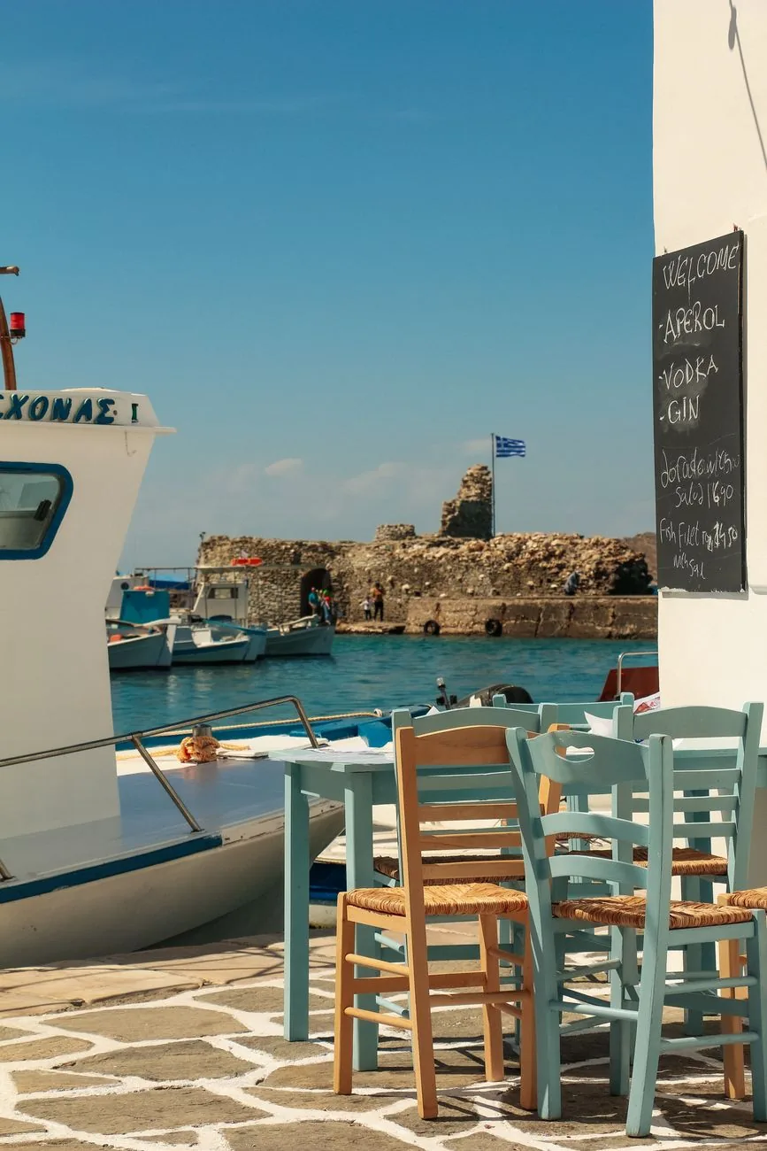 Tables et chaises paillées d'une taverne au bord d'un quai d'île grecque, avec un caïque amarré à quelques mètres