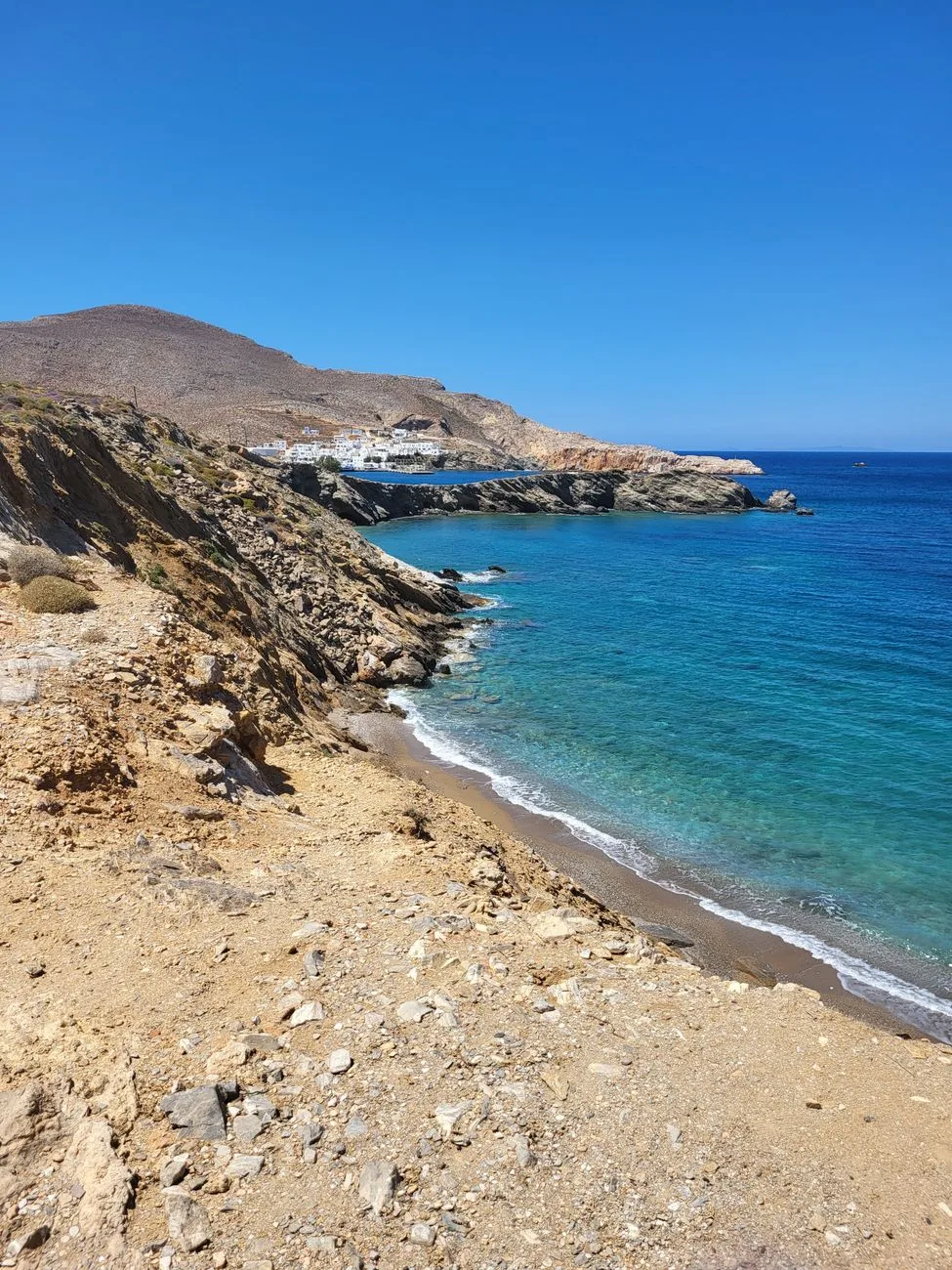 Plage de galets et de sable au pied des collines arides de Folegandros, avec les maisons blanches du port de Karavostasis visibles au fond de la côte