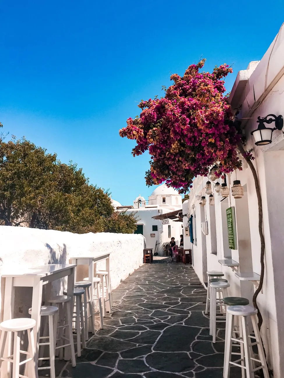 Ruelle dallée de la Chora de Folegandros bordée de murs blanchis, avec un bougainvillier en fleur et les tabourets d'un bar donnant sur une chapelle à dôme
