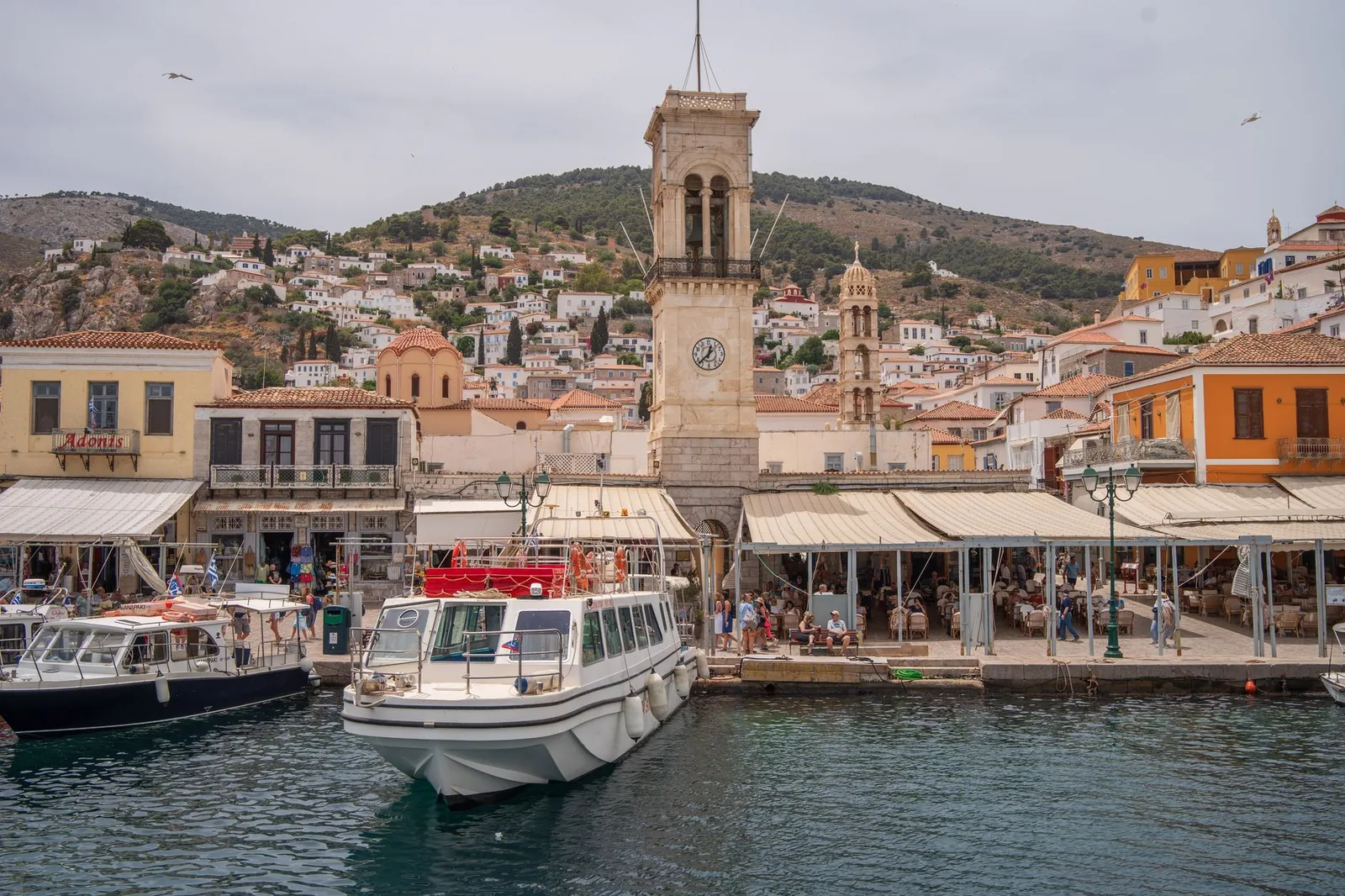 Le quai d'Hydra devant la tour de l'horloge du monastère de la Dormition, avec les bateaux amarrés et les terrasses de tavernes