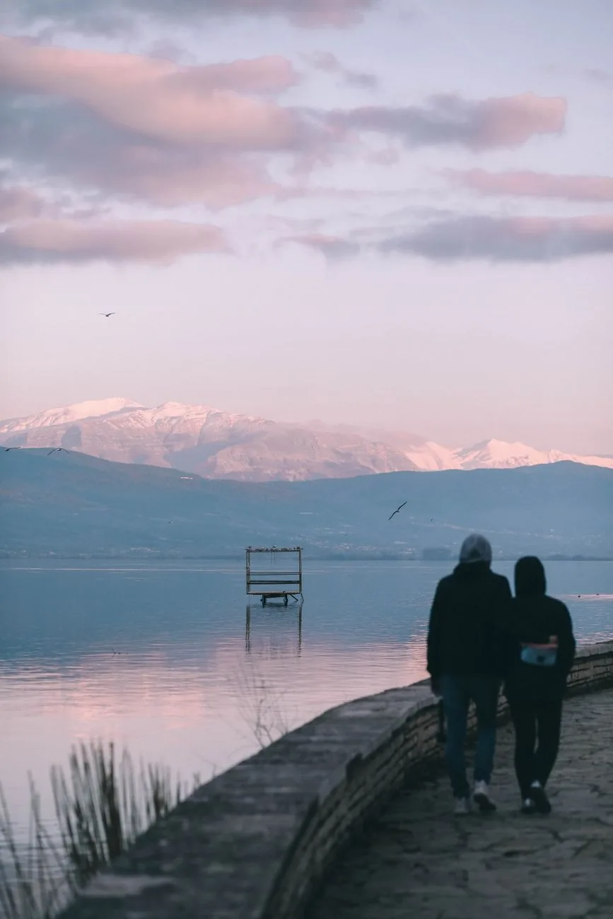 Un couple se promène sur la digue du lac d'Ioannina au coucher du soleil, avec les montagnes enneigées en face