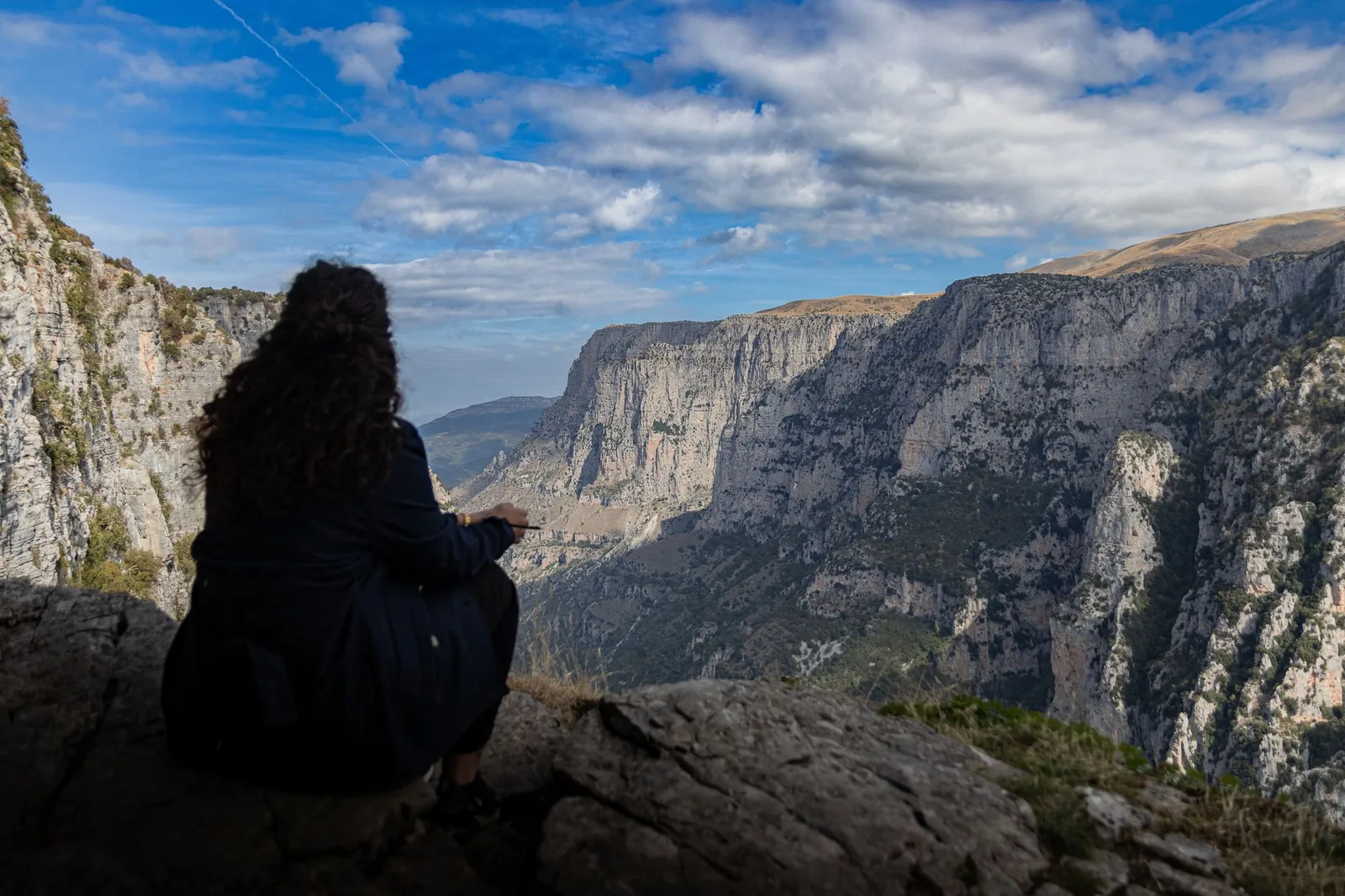 Une randonneuse assise sur un rocher au bord des gorges de Vikos, face aux falaises de calcaire de l'autre rive