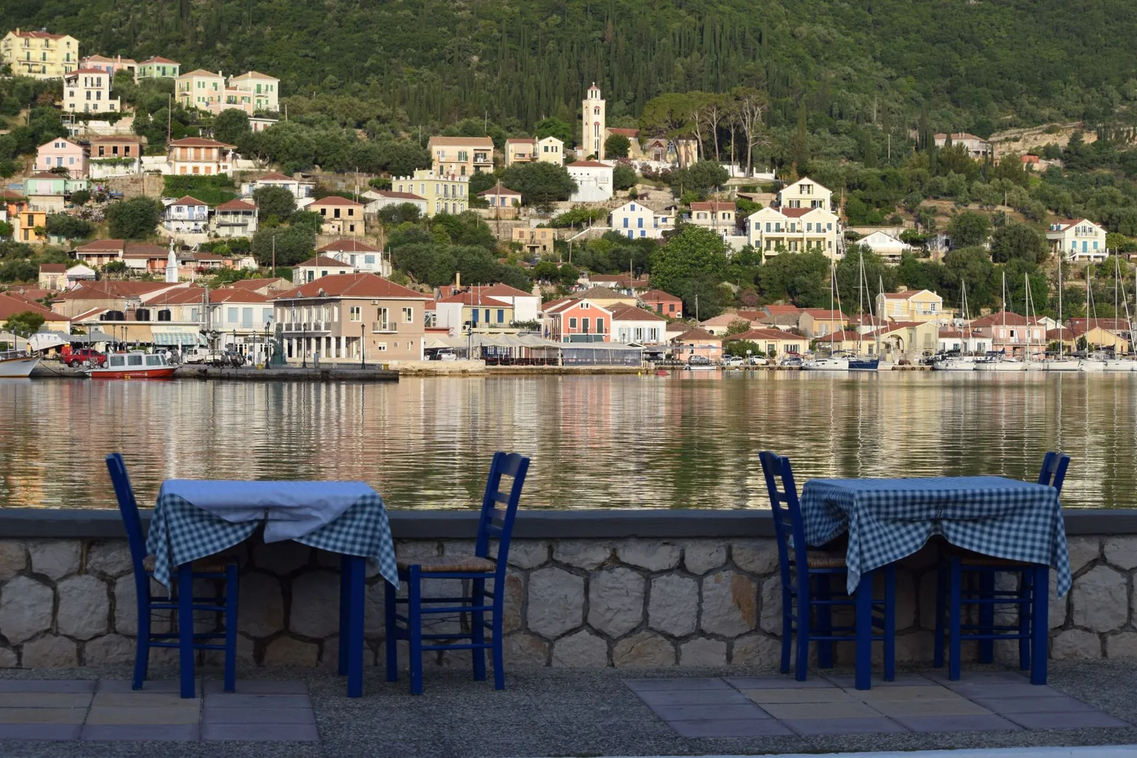 Terrasse de taverne aux chaises bleues face au port paisible et verdoyant de Vathy, à Ithaque