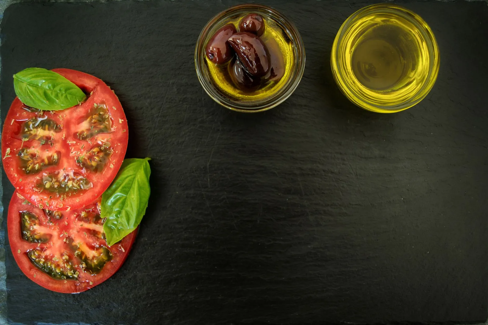 Olives noires de Kalamata en forme d'amande dans un bol d'huile et un verre d'huile d'olive extra vierge, avec tomates et basilic, sur une ardoise