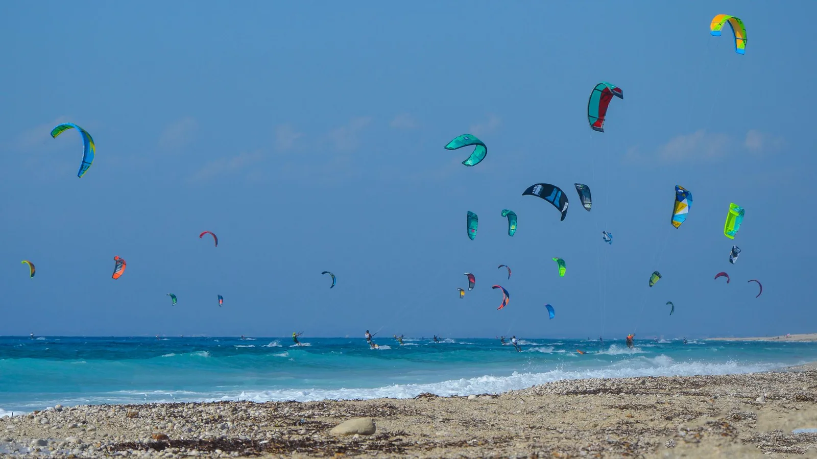 Dizaines d'ailes de kitesurf déployées au dessus d'une mer grecque balayée par le vent, vues depuis une plage de galets
