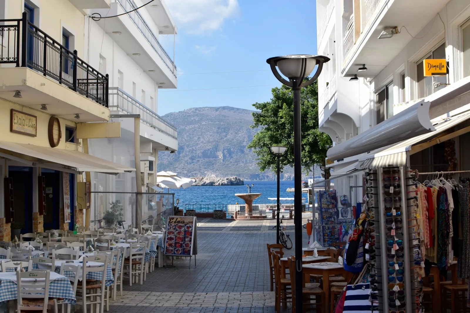 Rue piétonne de Pigadia à Karpathos, ses terrasses de tavernes et ses boutiques ouvrant sur la mer au bout de la perspective