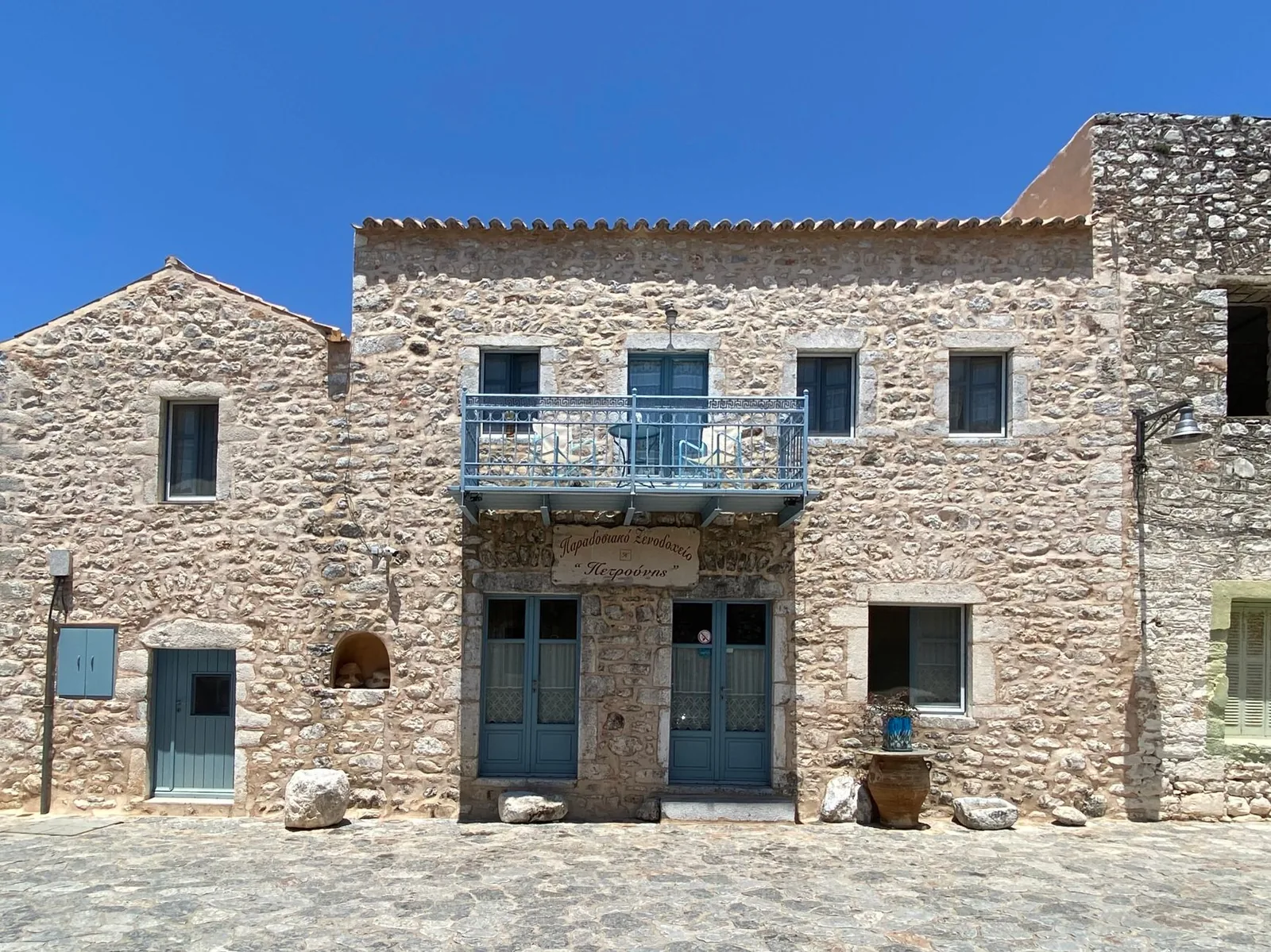 Façade en pierre d'un hôtel traditionnel du Magne avec ses volets bleu pâle et son balcon en fer forgé, sur une place pavée