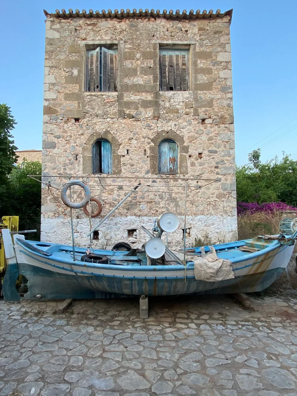 Maison-tour en pierre du Magne aux volets délavés, avec une barque de pêche bleue posée sur le pavage devant