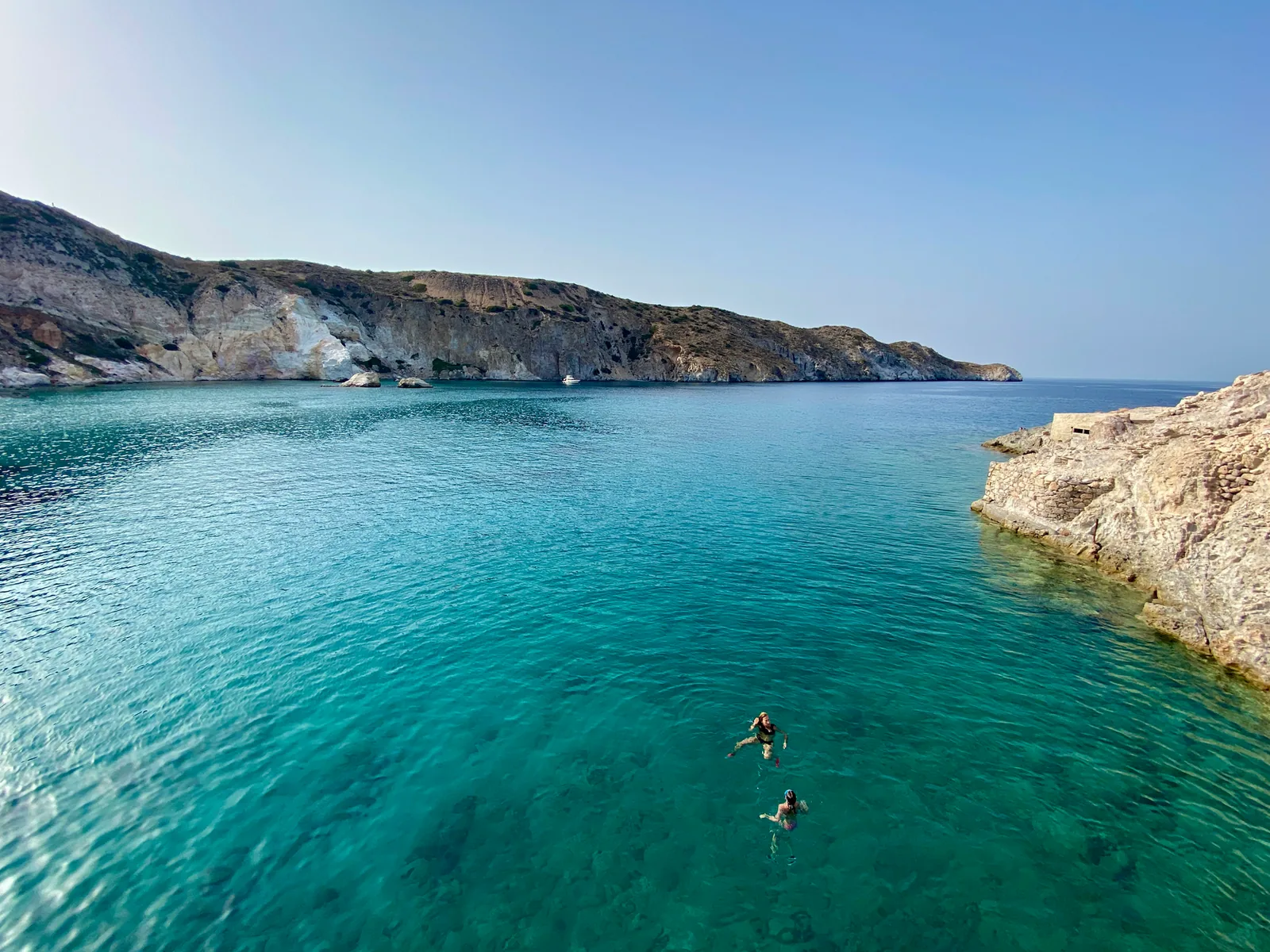 Baigneurs dans l'eau turquoise très claire de la baie de Firopotamos à Milos, au pied de falaises blanches et ocre surmontées de collines arides