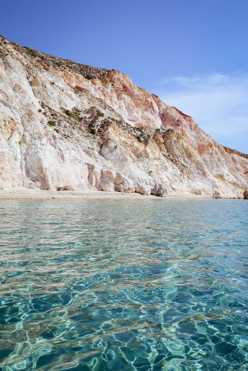 Falaises striées d'ocre, de rose et de blanc plongeant dans l'eau turquoise d'une plage de la côte sud de Milos