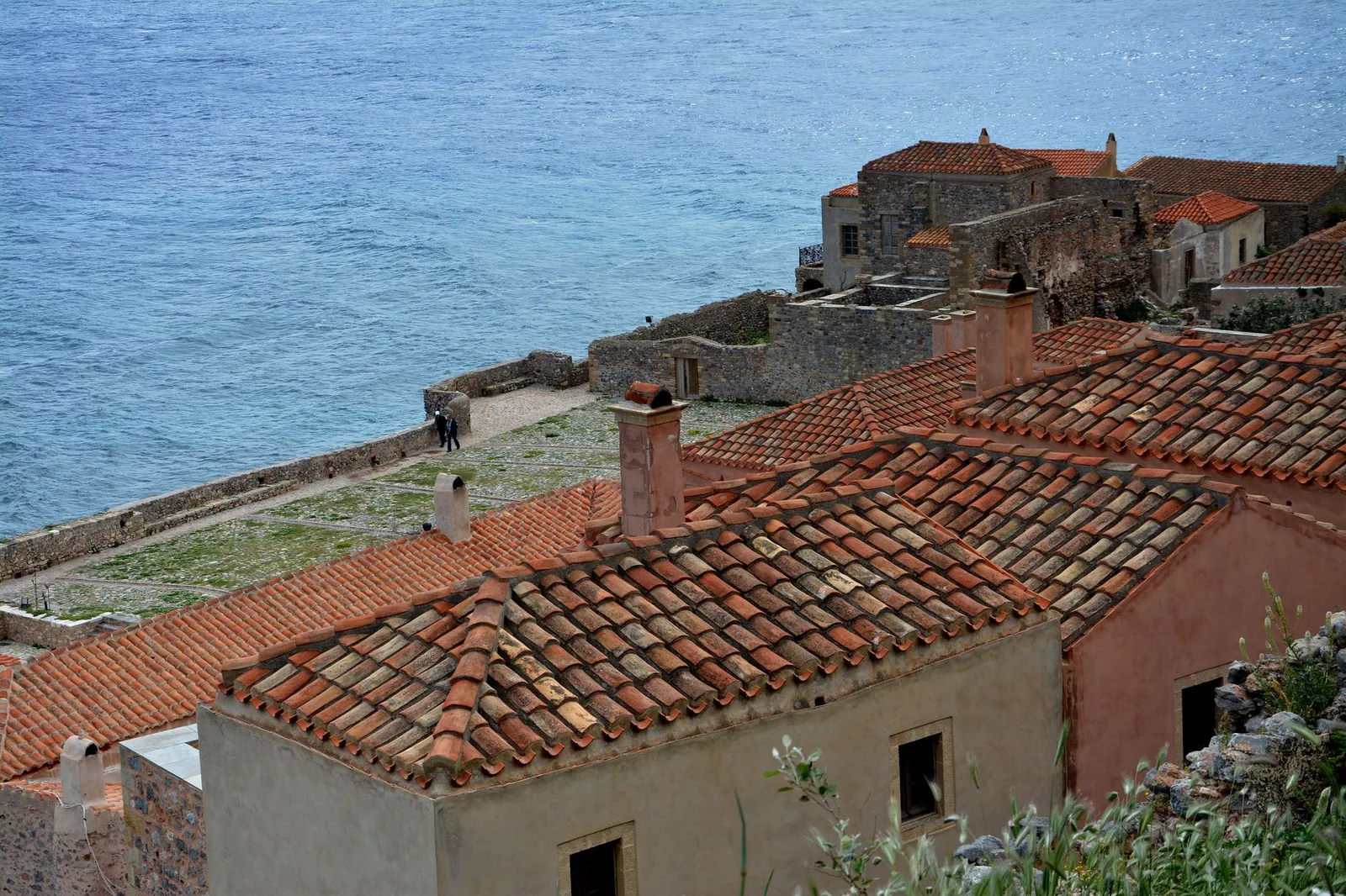 Deux promeneurs sur le chemin de ronde du rempart de Monemvasia, au-dessus de la mer, devant les toits de tuiles de la ville basse