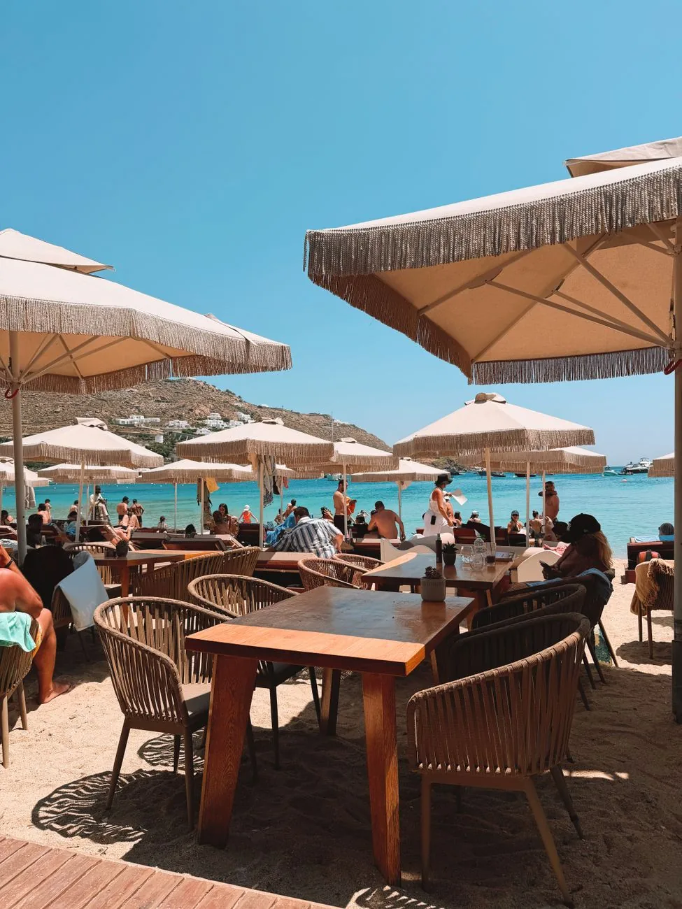 Beach club de Mykonos avec transats et parasols à franges face à une baie turquoise