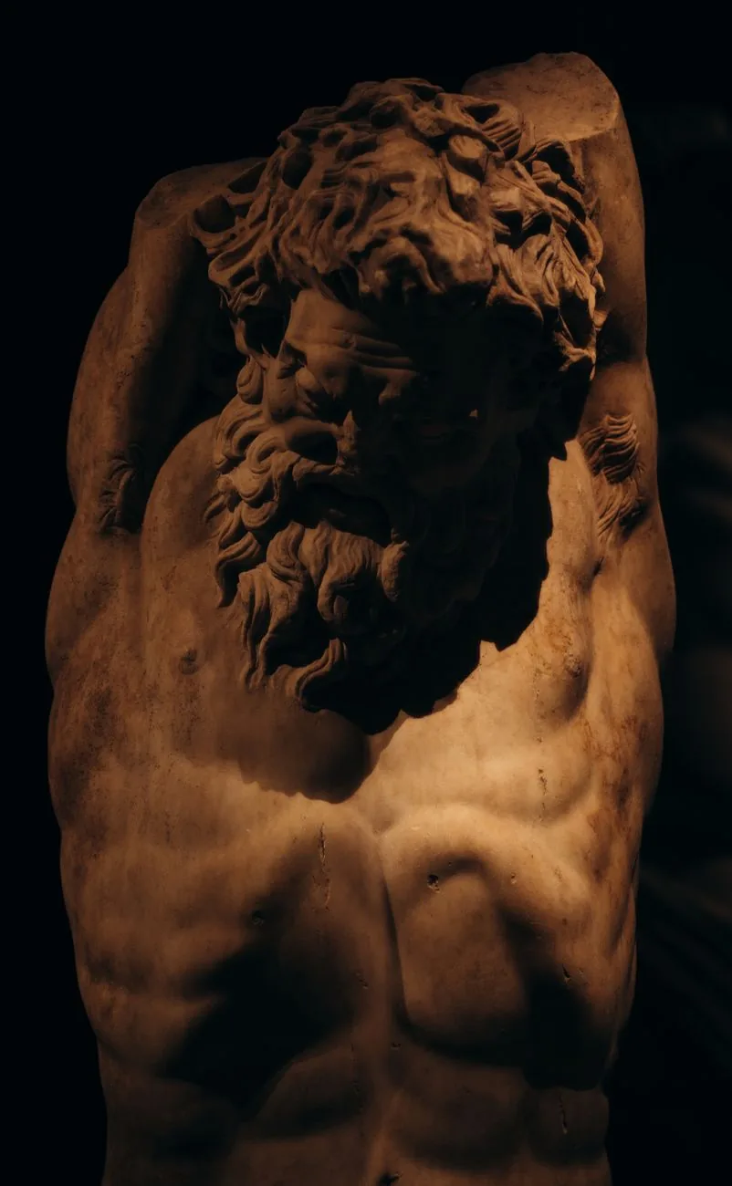Torse et visage barbu d'une statue grecque en marbre éclairée dans la pénombre, évoquant un Titan de la mythologie grecque