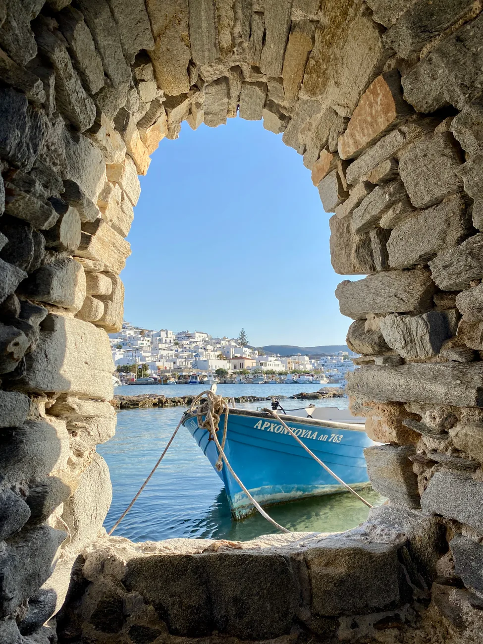 Vue à travers une arche de pierre du Kastro sur le port de Parikia et une barque de pêche bleue, à Paros