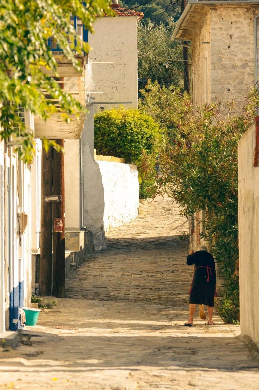 Ruelle pavée d'un village du Pélion en fin de matinée, bordée de maisons de pierre et de lauriers, une habitante en train de balayer