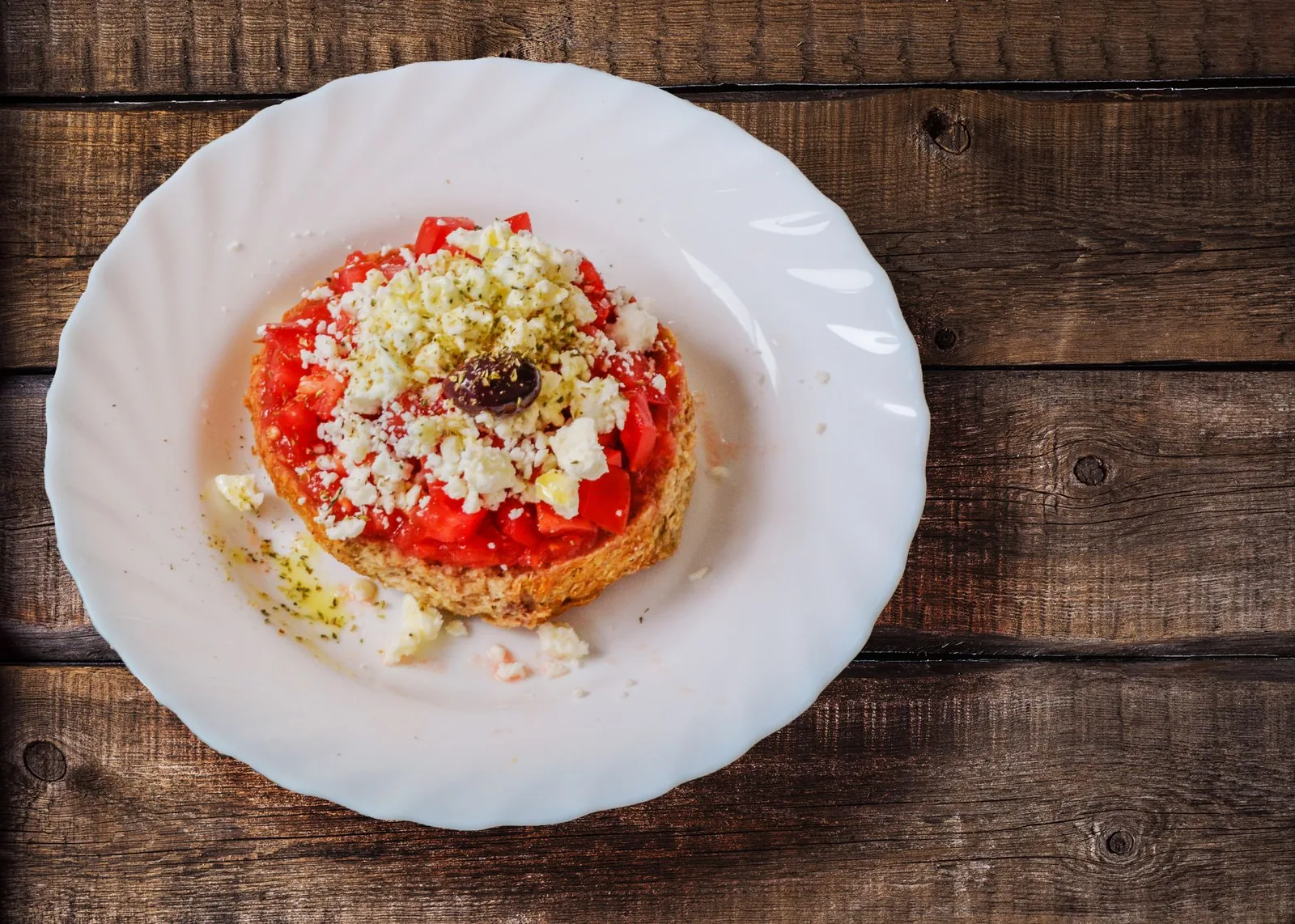 Dakos crétois servi dans une assiette blanche : galette d'orge ronde couverte de tomate concassée, de fromage de brebis émietté, d'origan et d'une olive noire