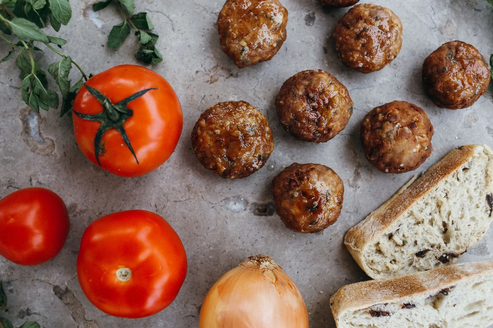 Keftedes grecs, boulettes de viande frites et dorées, posées sur une pierre claire avec des tomates, un oignon, de la menthe fraîche et du pain aux olives