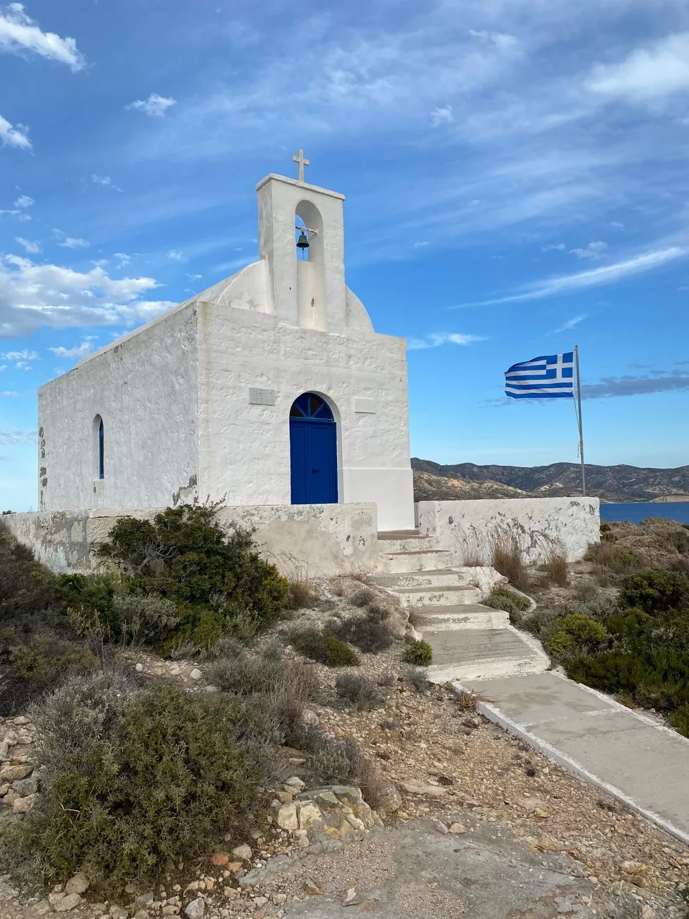 Chapelle blanche à porte bleue sur une colline de Sérifos, au bout d'un chemin bordé de broussailles, avec un drapeau grec et la mer en contrebas