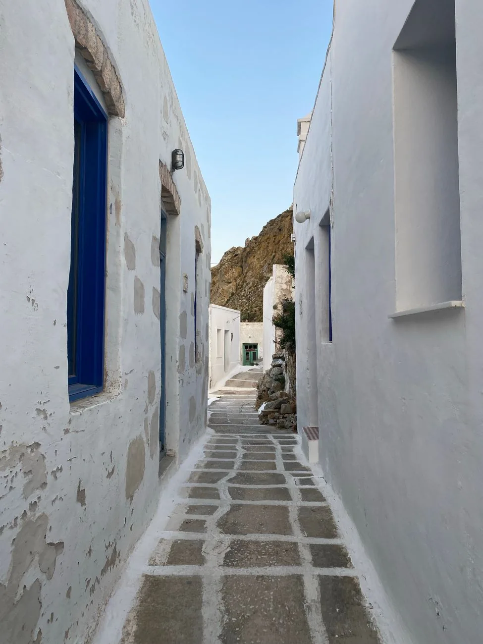 Ruelle dallée entre deux murs blanchis à la chaux dans la Chora de Sérifos, avec une fenêtre bleue et la roche de la colline au fond