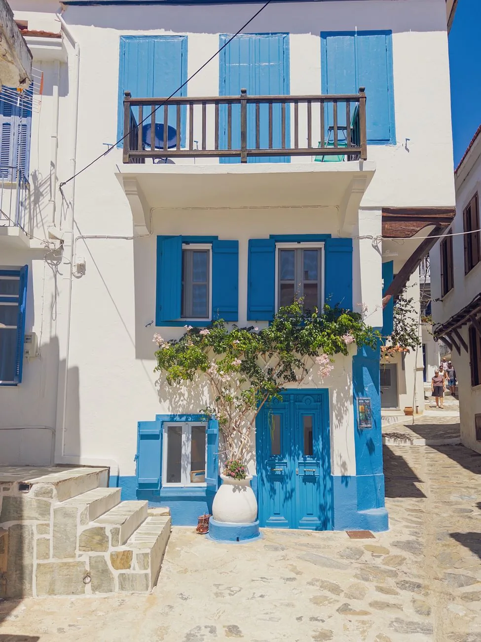 Maison blanche aux volets et balcon bleus dans une ruelle dallée de Skopelos Ville, avec une vigne sur la façade