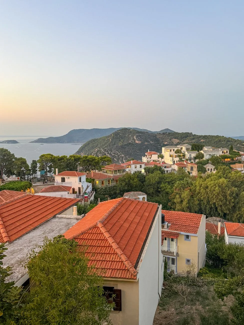 Toits de tuiles orange sur les hauteurs de Skopelos au coucher du soleil, avec la mer Égée et les collines boisées au loin