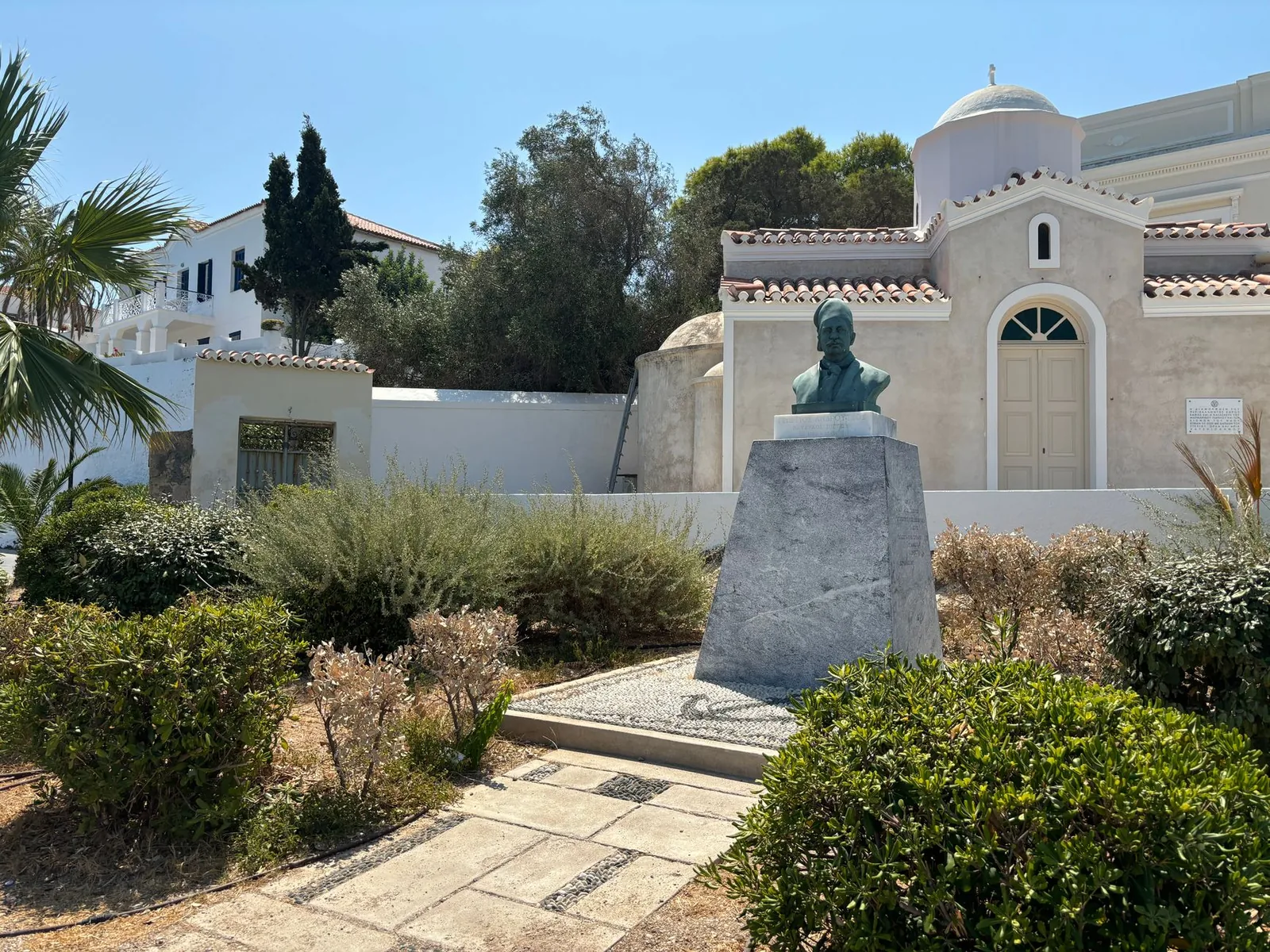 Buste en bronze sur un socle de pierre devant une petite église aux murs clairs, dans un jardin de Spetses