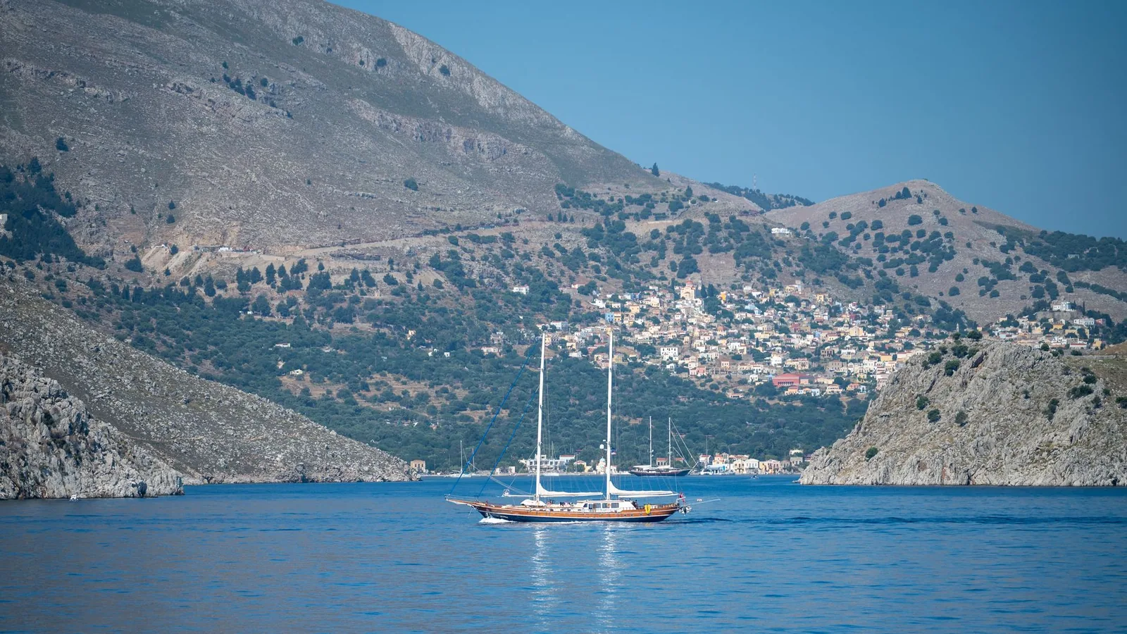 Voilier traditionnel à l'entrée de la baie de Symi, entre deux versants de collines sèches, le village étagé au fond