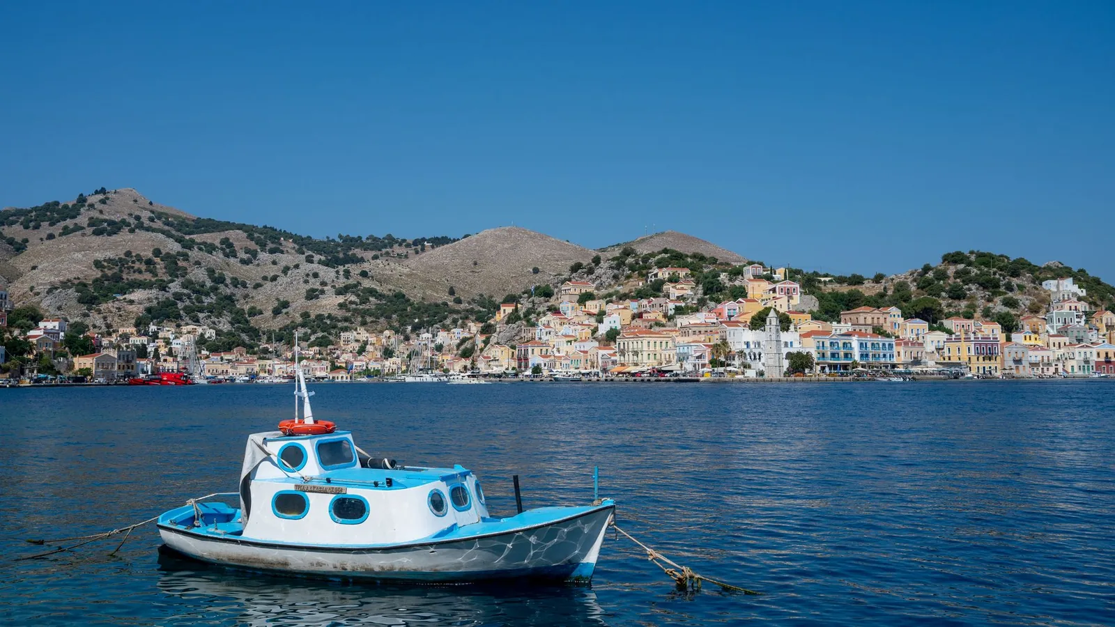 Petit bateau blanc et bleu amarré dans l'eau claire devant le front de mer coloré de Symi, du type de ceux qui desservent les criques