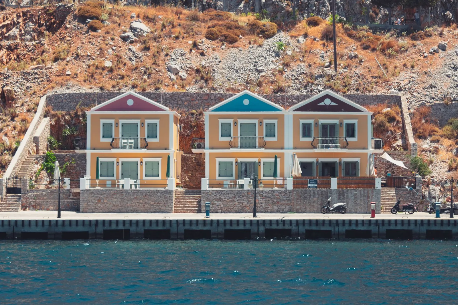 Trois maisons néoclassiques ocre à frontons colorés alignées sur le quai désert de Symi, au pied d'un versant rocheux