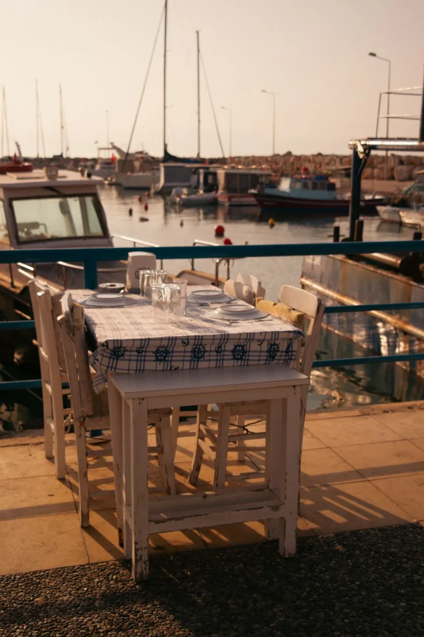 Table de taverne dressée sur le quai d'un port grec en fin de journée, nappe à motifs marins et barques amarrées derrière