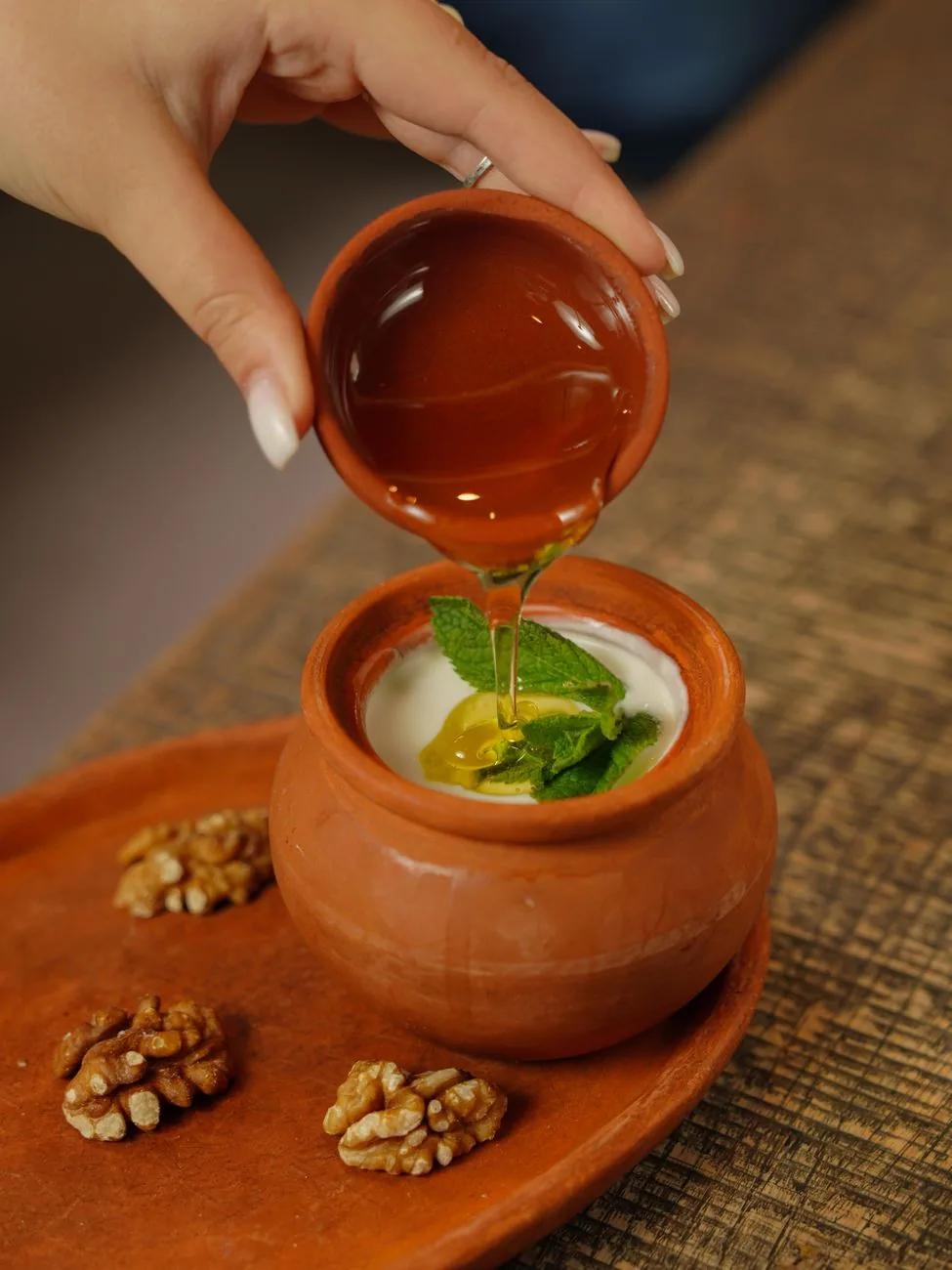Yaourt de brebis servi dans un pot en terre cuite, arrosé de miel versé d'une coupelle, avec des cerneaux de noix sur un plateau